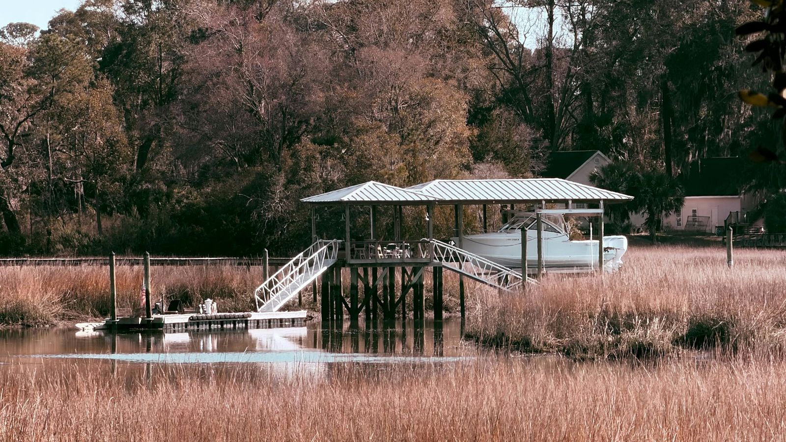 Covered private dock with a boat on a lift and stairs over golden salt marsh grass, trees behind
